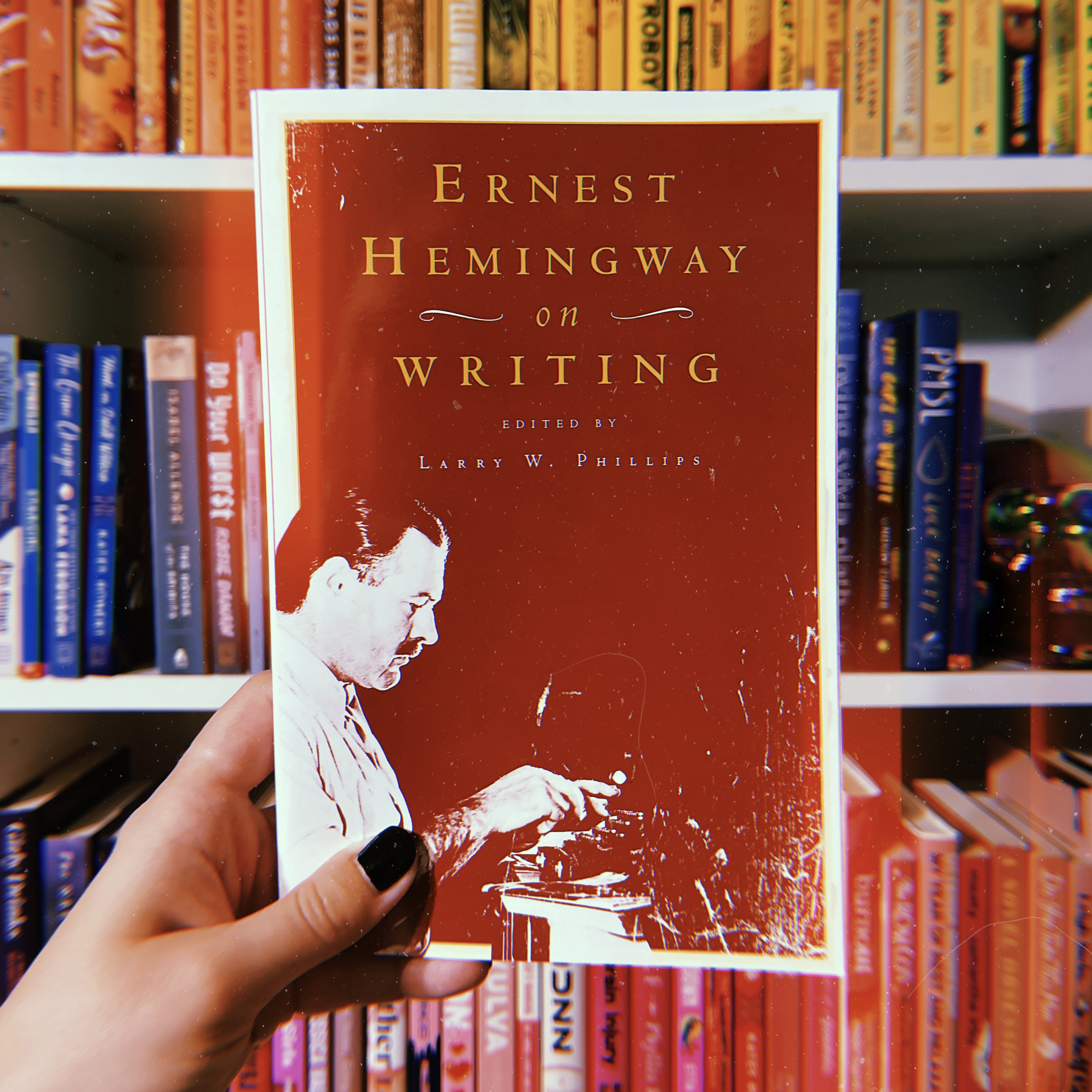 A white woman's hand with black fingernails holds a copy of Ernest Hemingway on Writing, edited by Larry W. Phillips.