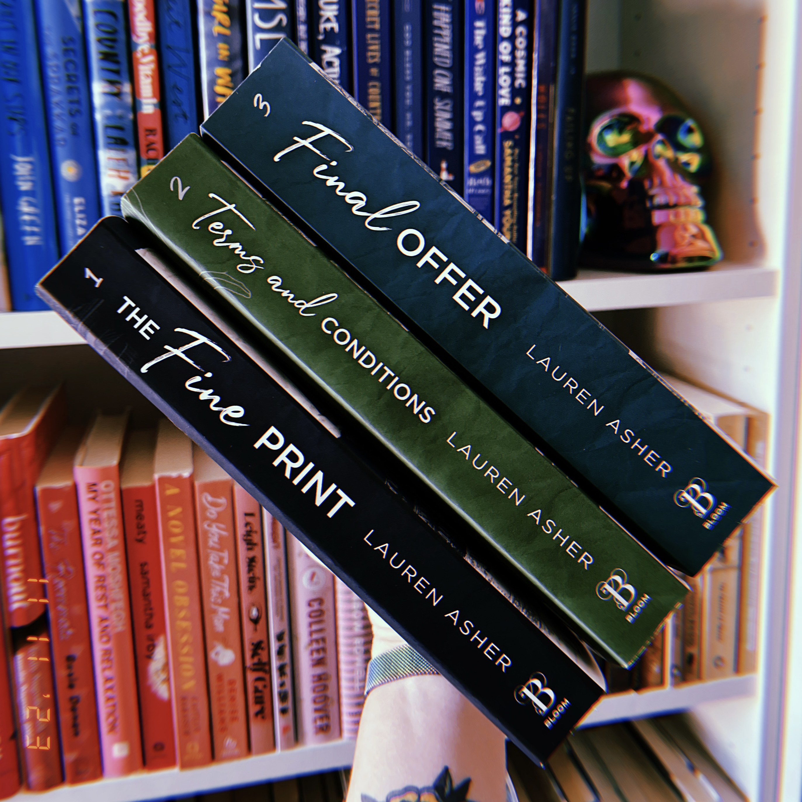 A hand holding a stack of books in front of a bookshelf. The books are The Fine Print, Terms and Conditions, and Final Offer, all by Lauren Asher.